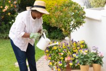 Donna anziana afroamericana che indossa guanti da giardinaggio sorridenti mentre innaffia le piante in giardino. rimanere in isolamento in isolamento in quarantena — Foto stock