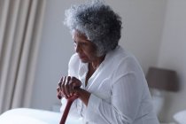 Thoughtful african american senior woman holding walking stick while sitting on bed at home. staying at home in self isolation in quarantine lockdown — Stock Photo