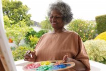 Femme âgée afro-américaine souriant tout en peignant sur toile debout sur le porche de la maison. rester en isolement personnel en quarantaine — Photo de stock