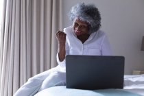 Femme âgée afro-américaine souriant tout en utilisant un ordinateur portable assis sur le lit à la maison. rester à la maison en isolement personnel en quarantaine — Photo de stock