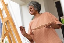 Mujer mayor afroamericana sonriendo mientras pinta sobre lienzo de pie en el porche de la casa. permaneciendo en aislamiento en cuarentena - foto de stock
