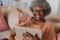 Femme âgée afro-américaine souriante et agitant tout en ayant un appel vidéo sur tablette numérique à la maison. rester à la maison en isolement personnel en quarantaine — Photo de stock