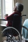Femme âgée afro-américaine réfléchie tenant une tasse de café regardant par la fenêtre alors qu'elle était assise sur un fauteuil roulant à la maison. rester à la maison en isolement personnel en quarantaine — Photo de stock