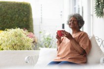 Pensativo afroamericano anciano mujer sosteniendo taza de café sonriendo mientras se sienta en el porche de la casa. permaneciendo en aislamiento en cuarentena - foto de stock