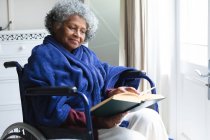 Mujer mayor afroamericana leyendo un libro mientras está sentada en silla de ruedas en casa. permaneciendo en casa en aislamiento en cuarentena - foto de stock