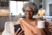 Eine afroamerikanische Seniorin hält eine Kaffeetasse lächelnd in der Hand, während sie zu Hause ein Videotelefon auf dem Smartphone führt. Zu Hause bleiben in Selbstisolierung in Quarantäne — Stockfoto
