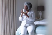 Pensativo afroamericano anciano mujer bebiendo café mientras está sentado en la cama en casa. permaneciendo en casa en aislamiento en cuarentena - foto de stock