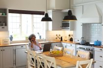 Mujer mayor afroamericana hablando por teléfono inteligente en la cocina en casa. permaneciendo en casa en aislamiento en cuarentena - foto de stock