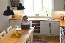 Africano americano sênior mulher falando no smartphone e usando laptop na cozinha em casa. ficar em casa em auto-isolamento em quarentena — Fotografia de Stock