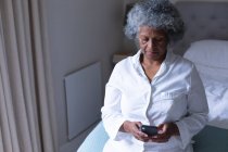 Thoughtful african american senior woman using smartphone while sitting on bed at home. staying at home in self isolation in quarantine lockdown — Stock Photo