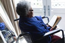 Eine afroamerikanische Seniorin liest ein Buch, während sie zu Hause im Rollstuhl sitzt. Zu Hause bleiben in Selbstisolierung in Quarantäne — Stockfoto