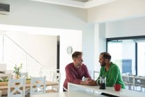 Casal masculino gay multi étnico sentado na cozinha bebendo café e falando em casa. Ficar em casa em auto-isolamento durante o bloqueio de quarentena. — Fotografia de Stock