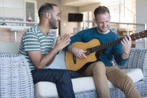 Multi ethnic gay male couple sitting on couch one playing guitar. staying at home in self isolation during quarantine lockdown. — Stock Photo
