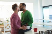 Couple masculin gay multi ethnique assis dans la cuisine buvant du café et s'embrassant à la maison. Rester à la maison en isolement personnel pendant le confinement en quarantaine. — Photo de stock
