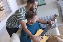 Multi pareja masculina gay étnica sentada en el sofá uno tocando la guitarra. permanecer en casa en aislamiento durante el bloqueo de cuarentena. - foto de stock