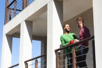 Couple masculin gay multi ethnique souriant et prenant un café sur le balcon au soleil. Rester à la maison en isolement personnel pendant le confinement en quarantaine. — Photo de stock