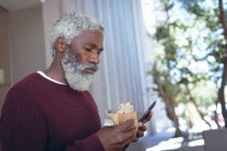 Ein afroamerikanischer Senior isst auf der Straße Sandwich und benutzt ein Smartphone. digitaler Nomade in der Stadt unterwegs. — Stockfoto