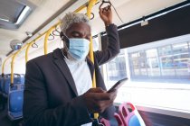 Африканский пожилой человек в маске для лица и наушниках, стоящих в автобусе со смартфоном. Цифровой кочевник в городе во время пандемии коронавируса. — стоковое фото