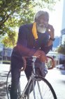 Homme âgé afro-américain portant un masque facial appuyé sur le vélo dans la rue parler sur smartphone. numérique nomade dehors et environ dans la ville pendant coronavirus covid 19 pandémie. — Photo de stock