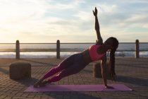 Mulher afro-americana exercitando-se no calçadão junto ao mar fazendo ioga. fitness estilo de vida ao ar livre saudável. — Fotografia de Stock