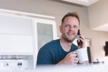 Hombre caucásico sonriendo, tomando café y hablando con un smartphone en casa. Permanecer en casa en aislamiento durante el bloqueo de cuarentena. - foto de stock