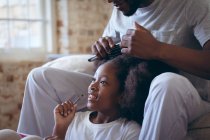 Homem afro-americano sentado na cama a pentear a filha. ficar em casa em auto-isolamento durante o confinamento de quarentena. — Fotografia de Stock