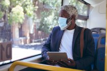 Hombre mayor afroamericano con máscara facial sentado en el autobús usando una tableta digital mirando por la ventana. nómada digital en la ciudad durante la pandemia de coronavirus covid 19. - foto de stock