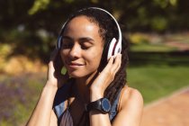 Smiling african american woman wearing headphones standing on scooter listening to music in street. Digital nomad on the go lifestyle. — Stock Photo