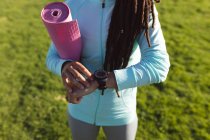 Средняя часть африканской американки тренируется в парке с помощью умных часов и ковриков для йоги. Здоровый образ жизни на открытом воздухе. — стоковое фото