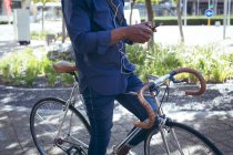 Низька частина чоловіка в навушниках сидить на велосипеді на вулиці, використовуючи смартфон. цифровий кочівник і близько в місті . — стокове фото