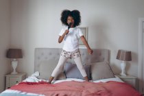 African american girl standing on bed holding hairbrush pretending to sing. staying at home in self isolation during quarantine lockdown. — Stock Photo