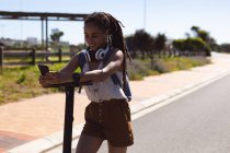 Sorrindo afro-americana usando fones de ouvido de pé na scooter usando smartphone na rua. Nômade digital em movimento estilo de vida. — Fotografia de Stock