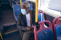 Hombre mayor afroamericano con máscara facial y auriculares sentados en el autobús sosteniendo el teléfono inteligente. nómada digital en la ciudad durante la pandemia de coronavirus covid 19. - foto de stock
