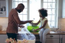 Chica afroamericana y su padre clasificando el reciclaje juntos en la cocina. permanecer en casa en aislamiento durante el bloqueo de cuarentena. - foto de stock