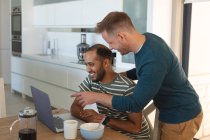 Multi pareja gay masculina étnica sonriendo y desayunando y usando el portátil en casa. Permanecer en casa en aislamiento durante el bloqueo de cuarentena. - foto de stock