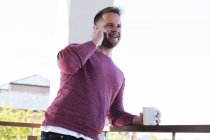 Smiling caucasian man holding cup of coffee and talking on smartphone on balcony in sun. Staying at home in self isolation during quarantine lockdown. — Stock Photo