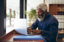 Afro-americano idoso sentado à mesa no café a trabalhar com laptop. nômade digital para fora e sobre na cidade. — Fotografia de Stock