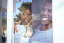 La ragazza afroamericana e suo padre tengono in braccio un orsacchiotto che guarda attraverso la finestra. stare a casa in isolamento durante l'isolamento in quarantena. — Foto stock