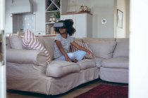Afroamerikanerin sitzt im Wohnzimmer mit vr headset. Während der Quarantäne zu Hause bleiben und sich selbst isolieren. — Stockfoto