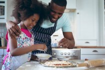 Chica afroamericana y su padre haciendo pizza juntos en la cocina. permanecer en casa en aislamiento durante el bloqueo de cuarentena. - foto de stock