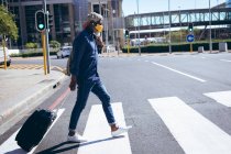 Hombre mayor afroamericano usando mascarilla facial y auriculares con maleta de ruedas cruzando la calle en el cruce peatonal. nómada digital en la ciudad durante la pandemia de coronavirus covid 19. - foto de stock