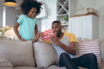 Menina afro-americana de pé na sala de estar dando um presente ao pai. ficar em casa em auto-isolamento durante o confinamento de quarentena. — Fotografia de Stock