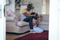 African american man and his daughter sitting in living room using vr headset. staying at home in self isolation during quarantine lockdown. — Stock Photo