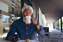 Ein älterer afroamerikanischer Mann sitzt am Tisch vor einem Café, trinkt Kaffee mit seinem Smartphone und schaut weg. digitaler Nomade in der Stadt unterwegs. — Stockfoto