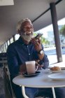 Hombre mayor afroamericano sentado en la mesa fuera de la cafetería con café hablando en el teléfono inteligente y sonriendo. nómada digital en la ciudad. - foto de stock