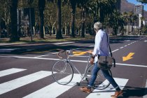 African american senior man wheeling bicycle across road on a pedestrian crossing. digital nomad out and about in the city. — Stock Photo