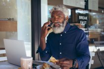 Hombre mayor afroamericano sentado en la mesa fuera de la cafetería hablando en el teléfono inteligente. nómada digital en la ciudad. - foto de stock