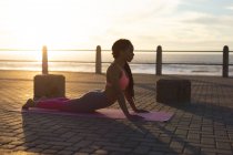 Mujer afroamericana haciendo ejercicio en el paseo marítimo haciendo yoga. fitness estilo de vida saludable al aire libre. - foto de stock