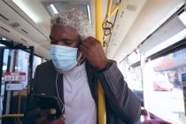Hombre mayor afroamericano con máscara facial que se pone auriculares de pie en el autobús usando un teléfono inteligente. nómada digital en la ciudad durante la pandemia de coronavirus covid 19. - foto de stock