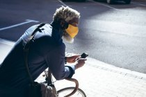 Hombre mayor afroamericano con máscara facial apoyada en bicicleta en la calle usando un teléfono inteligente. nómada digital en la ciudad durante la pandemia de coronavirus covid 19. - foto de stock